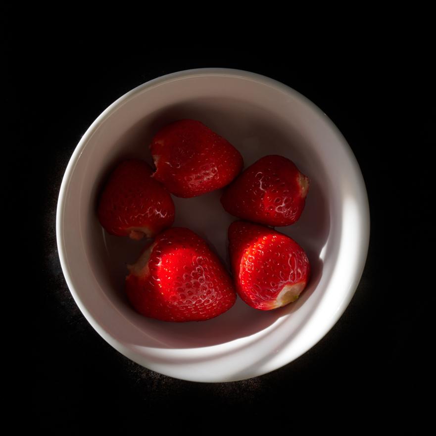 Five vibrant, glossy red strawberries in a white ceramic bowl, dramatically lit against a stark black background.