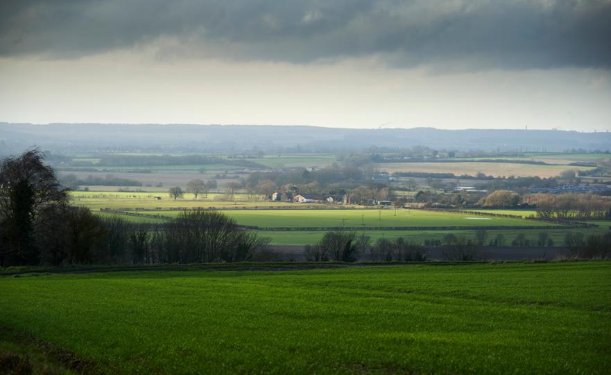 Expansive rural landscape with vibrant green fields, distant farm buildings, bare trees, and a dramatic, overcast sky.